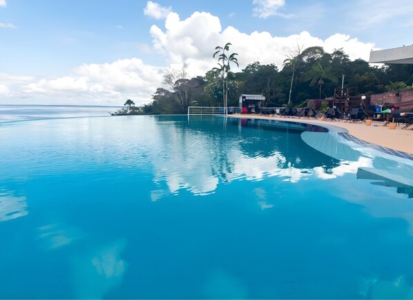 Una piscina azul cristalina con vista al océano, rodeada de palmeras y una zona de playa con sillas de descanso.