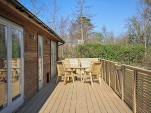 A wooden deck with a table and chairs, a glass door, and a view of trees and a hedge. - 4 Southern Place (Liskeard)