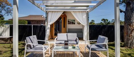 An outdoor patio with a pergola, two cushioned chairs, a small table, and a decorative item.