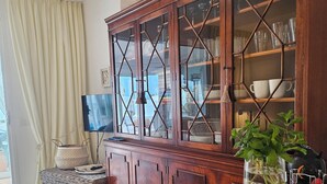 Uma sala de estar com televisão, um armário de madeira com portas de vidro, um banquinho de vime e uma mesa de jantar com uma tigela de frutas e um vaso de flores.