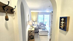 Corredor que dá para um quarto com cama, mesa de cabeceira, abajur e vista para o exterior.