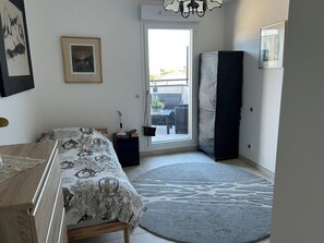 Un dormitorio con cama, mesita de noche, una silla, ventana y puerta que da a un balcón.