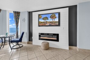 A modern living room with a fireplace, a television mounted above it, and a dining area with a view of the ocean. - Atlantic Resort 502 – Relaxing Oceanfront Getaway (Myrtle Beach)