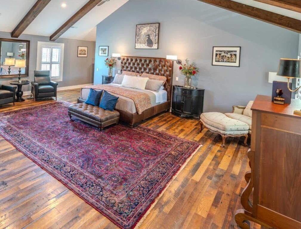 A bedroom with a large bed, a sitting area with a chair and ottoman, a nightstand, and a dresser.