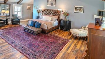 A bedroom with a large bed, a sitting area with a chair and ottoman, a nightstand, and a dresser.