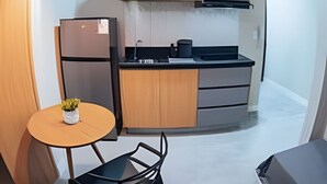 Cocina compacta con armarios de madera, microondas y refrigerador. Hay una mesita redonda con una silla y una planta en maceta.
