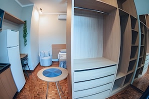 A compact room with a bed, a small table, a chair, a refrigerator, and a plant.