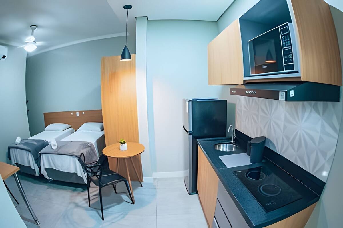 A compact hotel room with a bed, a small table with a chair, a kitchenette with a microwave and stove, and a ceiling fan.
