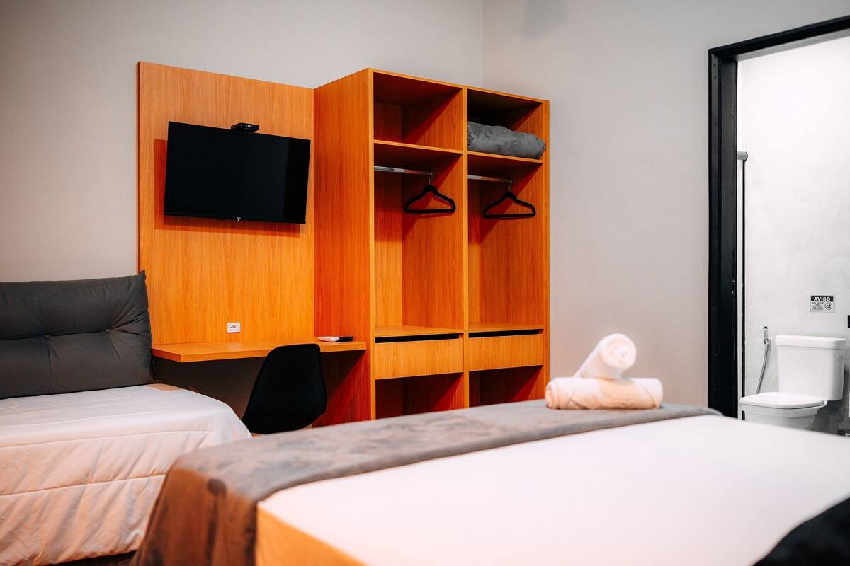 A room with a bed, a wooden wardrobe with a TV, a chair, and a toilet.