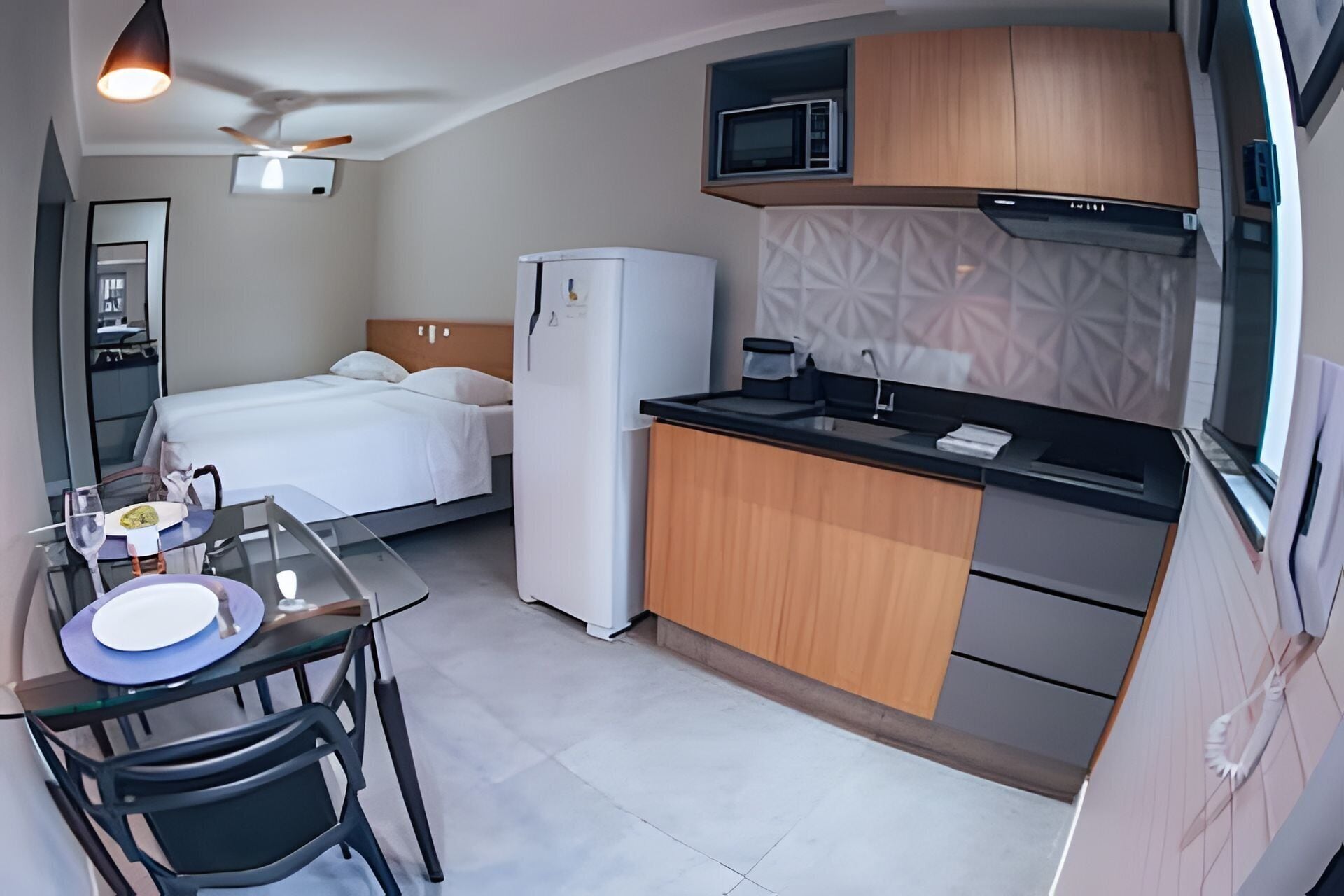 A compact hotel room with a kitchenette, a bed, a dining area, and a ceiling fan.