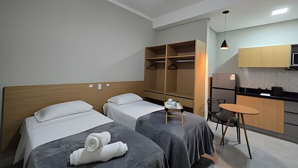 A hotel room with two beds, a small table, a chair, a kitchenette, and a closet. - Zuccaro Studio (Bauru)