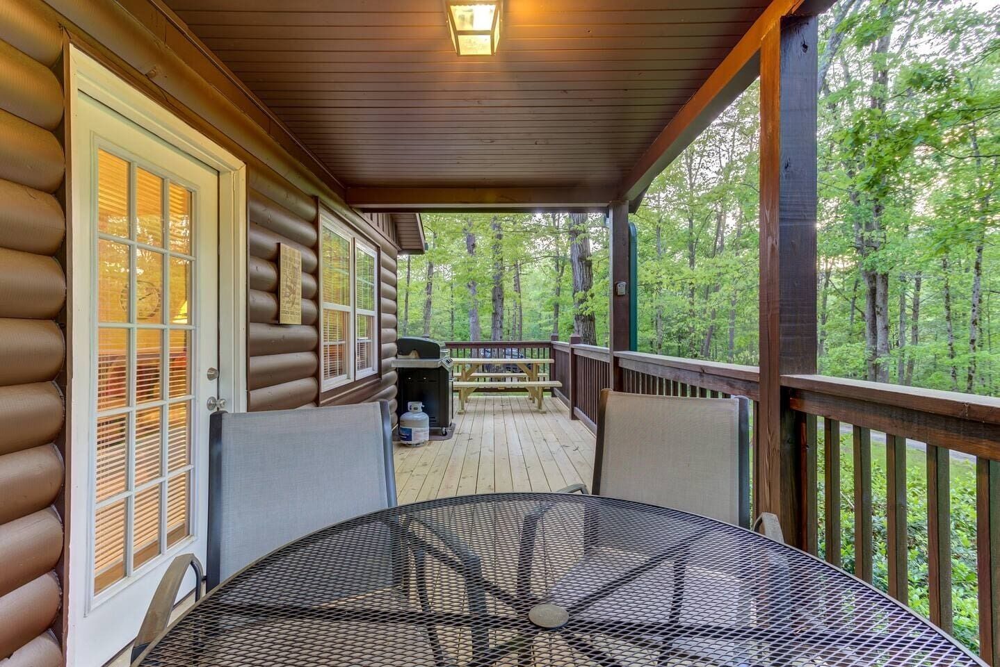 A covered outdoor deck with a metal table and benches, surrounded by wooden railings and a forested backdrop.