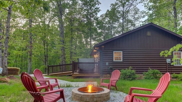 Una cabaña de troncos con una fogata y sillas Adirondack rojas en un entorno forestal.