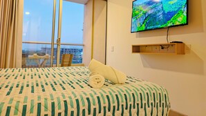 Habitación de hotel con cama, televisor montado en la pared, ventana con cortinas y balcón con mesa y sillas.