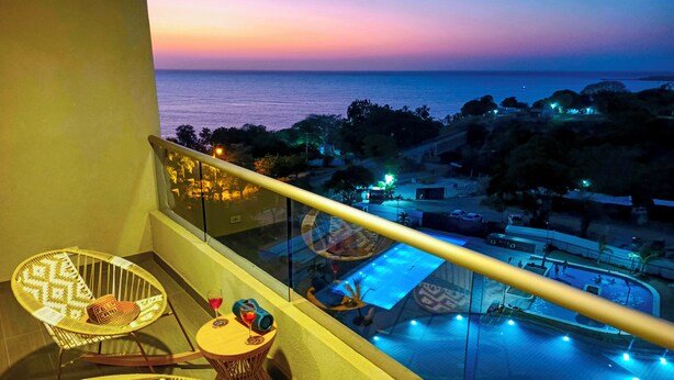 Un balcón con sillas de mimbre y una mesa pequeña, con vista a una zona de piscina y al atardecer.