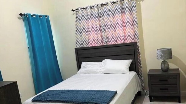 A bedroom with a bed, a nightstand, a lamp, a blue curtain, and a patterned rug. - Terrific 2-bedroom house with AC in Montego Bay (Montego Bay)