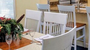 Un coin repas avec une table en bois, des chaises blanches et une cuisine en arrière-plan.