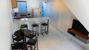 A compact kitchen with a refrigerator, a dining area with a round table and chairs, and a TV mounted on the wall. - Condomínio Triangulo da Embauba  (Porto Seguro)