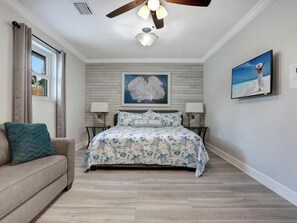 Ein Schlafzimmer mit einem Bett, einem Fenster mit Vorhängen, einem Deckenventilator, einem Wand-Fernseher und einem Gemälde.