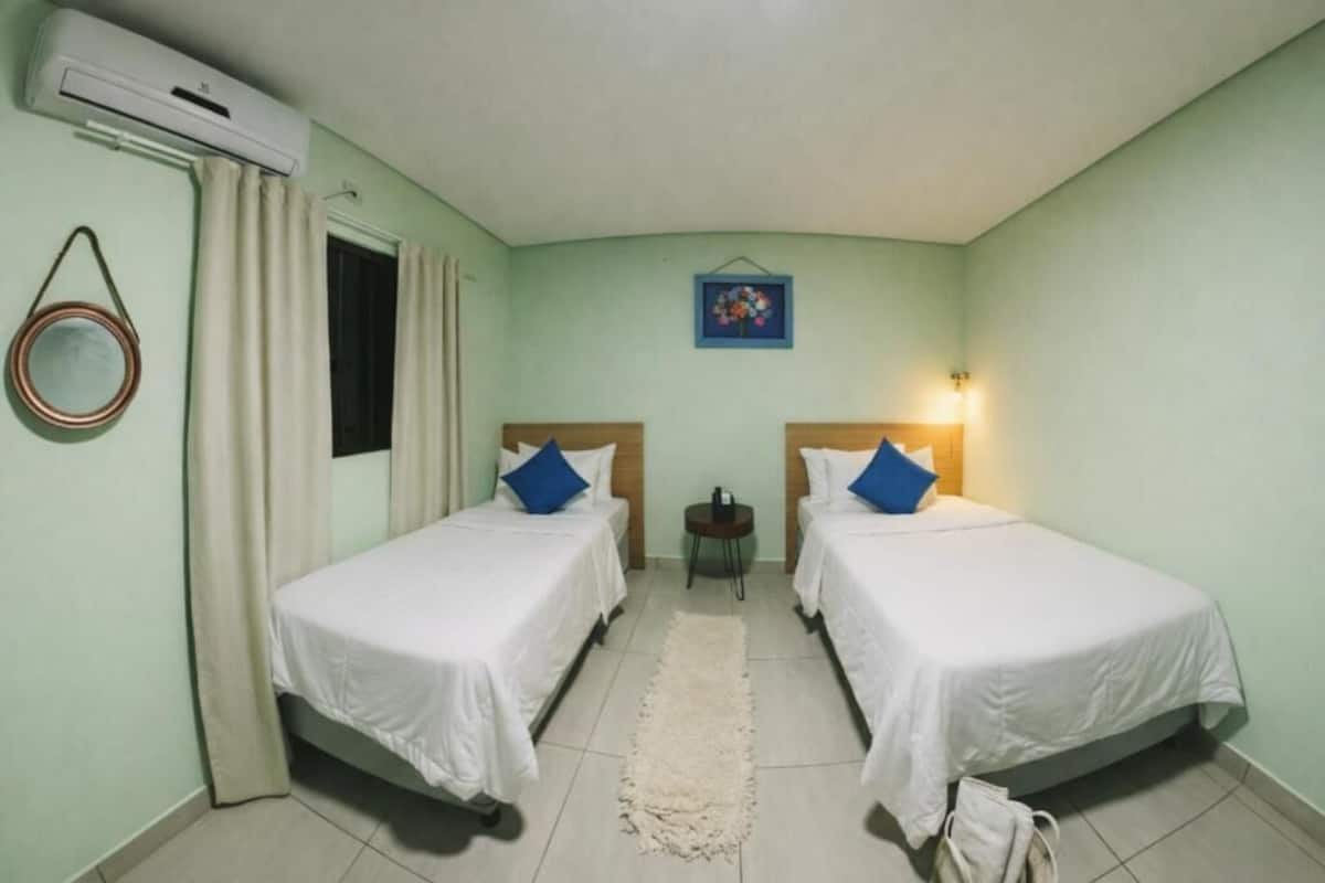 A room with two single beds, a small round table, a wall-mounted air conditioner, and a framed picture on the wall.