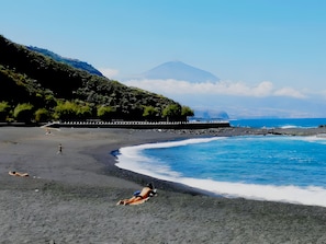 Une plage bordée d’une côte rocheuse, avec une montagne qui se profile à l’horizon et une jetée qui s’avance dans l’eau.