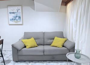A grey sofa with yellow cushions, a framed picture on the wall, a small table with a vase, and a window with curtains. - Sunset Beach Apartment Tenerife (Tacoronte)