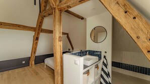 A bedroom with exposed wooden beams, a white bed, a bathroom with a blue-tiled shower, and a round mirror. - Manoir de charme en Normandie | piscine chauffée à moins de 2h de Paris. (Chambord)