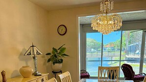A dining area with a glass-top table, white chairs, a chandelier, a clock, and a view of a body of water through large windows. - Charming 2-bedroom apartment with AC in perfect Naples (Naples)