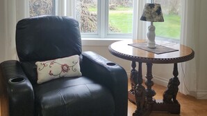 A room with a black leather armchair, a small round table with a lamp, and a view through large windows.