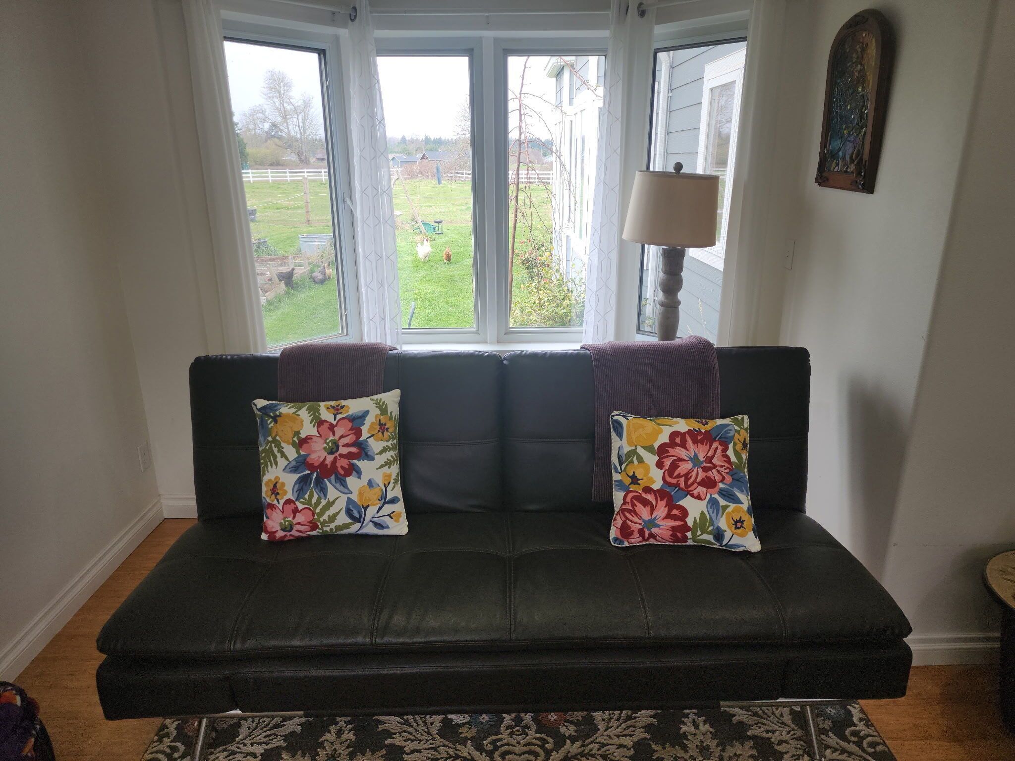 A dark leather sofa with floral cushions, a window with a view of a yard, and a lamp on a side table.