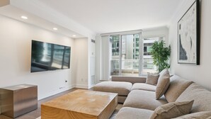 A modern living room with a flat-screen TV, a wooden coffee table, a beige sofa, and a large window with a view of a building. - Luxury DT Escape 3Bed2Bath! Pool |Gym |Parking (Toronto)