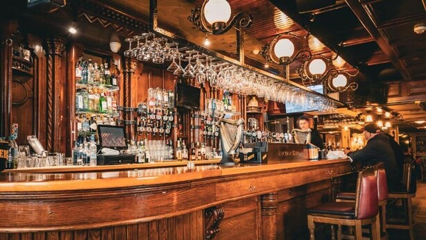 Un bar con rivestimenti in legno, diverse bottiglie, bicchieri e un barista al lavoro.
