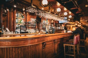 Um bar com painéis de madeira, diversos frascos, copos e uma pessoa trabalhando atrás do balcão.