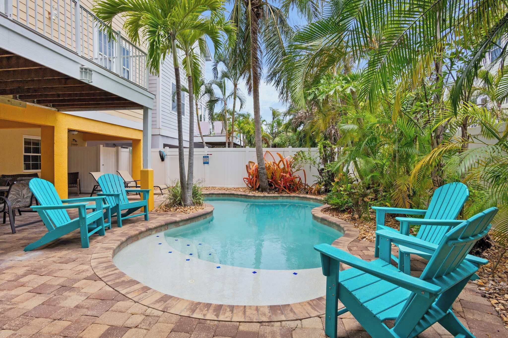 A swimming pool with turquoise Adirondack chairs, surrounded by palm trees and a covered patio area.