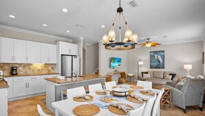 A modern kitchen with white cabinets, a central island, and a dining area with white chairs and a table set with placemats. - 5 Min Walk to Beach | Private Pool | AMI Trolley (Holmes Beach)