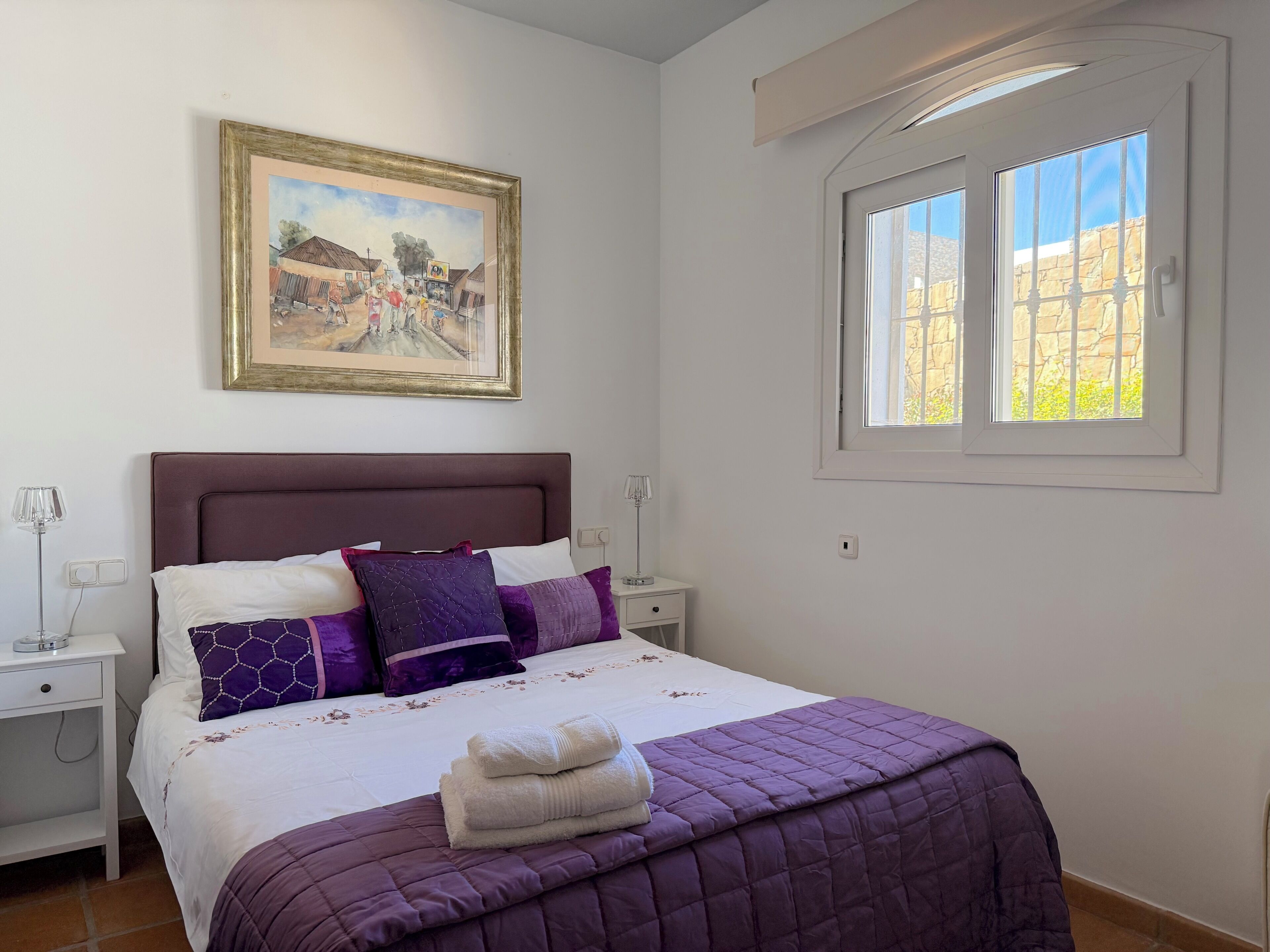 A neatly made bed with a purple quilt, a framed painting above it, and a window with a stone wall view.