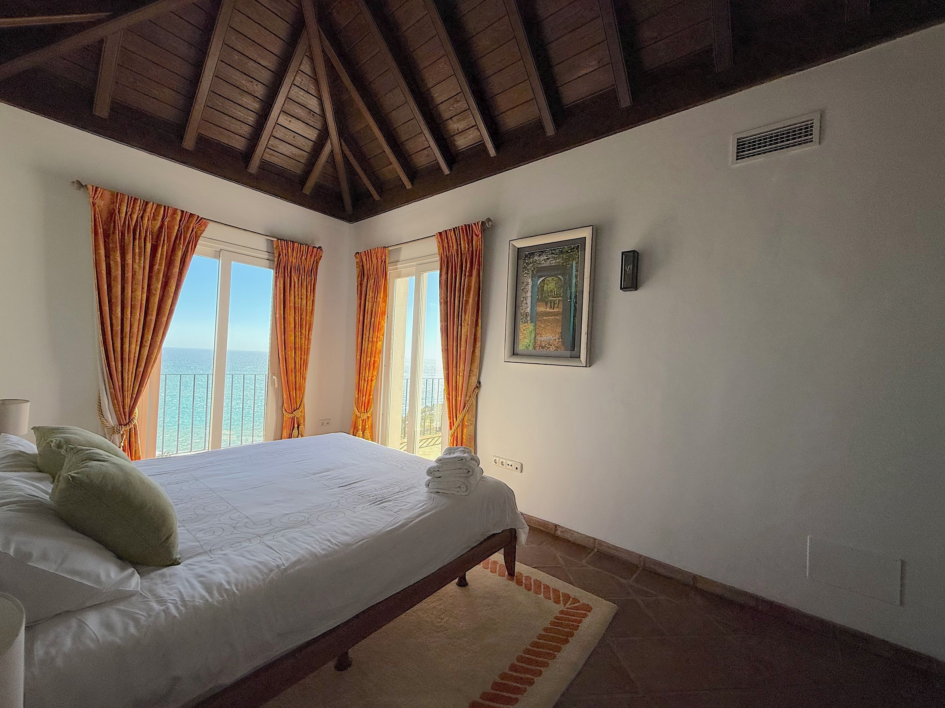 A bedroom with a bed, curtains, a picture on the wall, and a view of the sea.