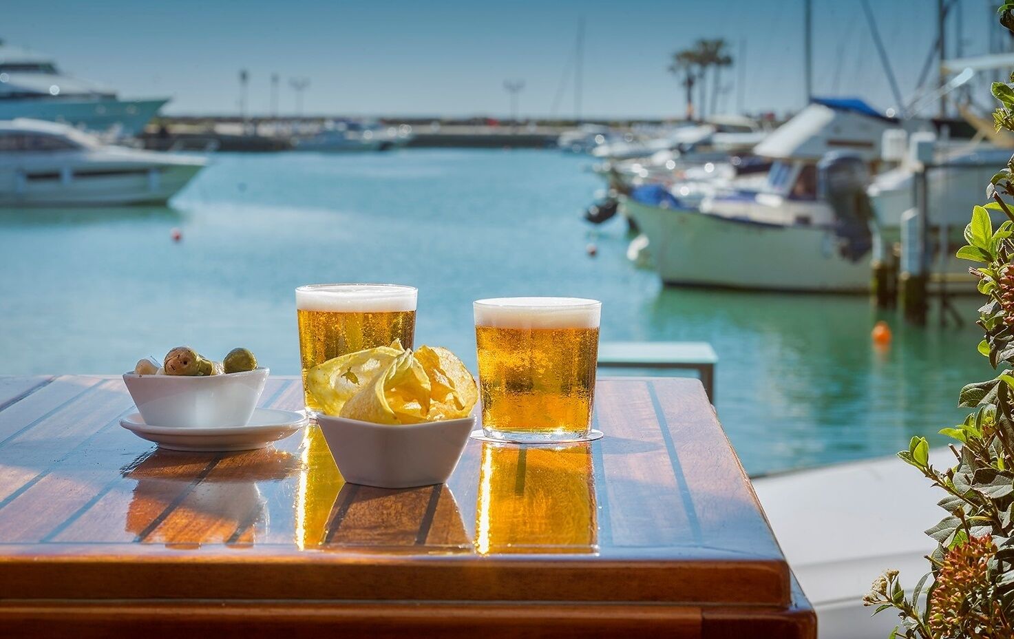 Une table avec deux verres de bière et un bol de collations, près d’un port de plaisance.