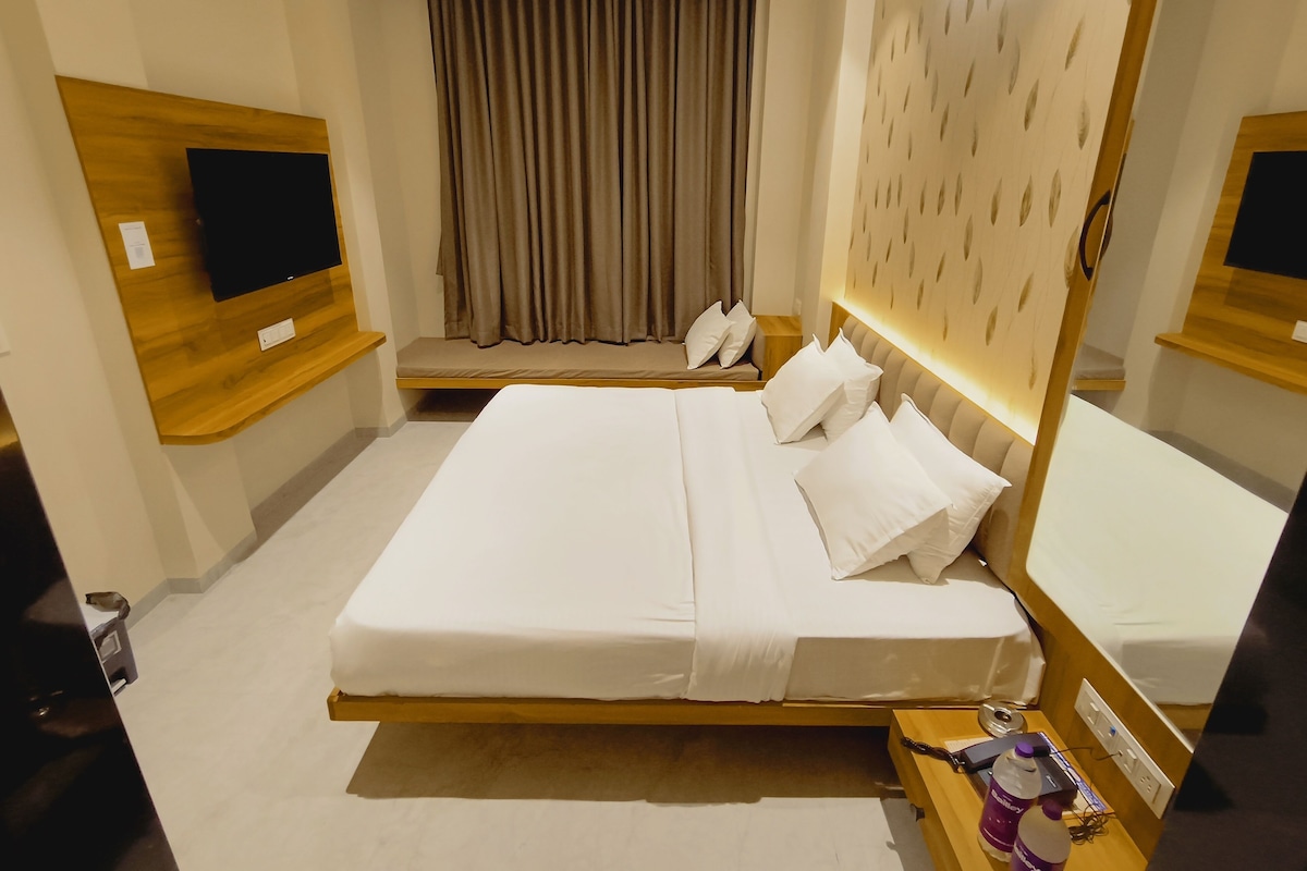 A hotel room with a large bed, a bedside table with a lamp, a TV mounted on the wall, and a window with curtains.