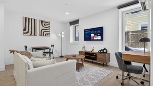 A modern living room with a white sofa, a wooden coffee table, a TV mounted on the wall, and a desk with a chair. - Luxury 2BR | Free Parking | Harvard/BU | ROKU TV (Boston)