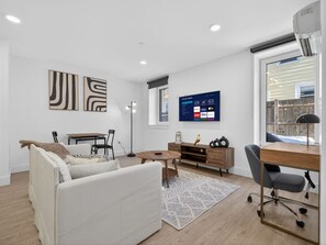Ein modernes Wohnzimmer mit einem weißen Sofa, einem Holztisch, einem an der Wand montierten Fernseher und einem Schreibtisch mit Stuhl.
