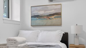 A neatly made bed with white linens, a gray throw blanket, and a stack of white towels. There is a painting on the wall above the bed, a bedside lamp on a wooden nightstand, and a window with blinds. - Luxury 2BR | Free Parking | Harvard/BU | ROKU TV (Boston)