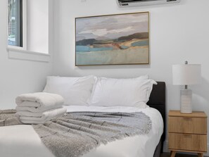 Ein ordentlich bezogenes Bett mit weißen Leinentextilien, einer grauen Tagesdecke und einem Stapel weißer Handtücher. Darüber an der Wand hängt ein Gemälde, auf dem Nachttisch steht eine Nachttischlampe und es gibt ein Fenster mit Jalousien.