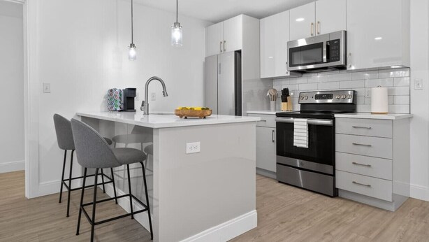 A modern kitchen with white cabinetry, a central island with bar stools, stainless steel appliances, and pendant lighting. - Luxury 2BR | Free Parking | Harvard/BU | ROKU TV (Boston)