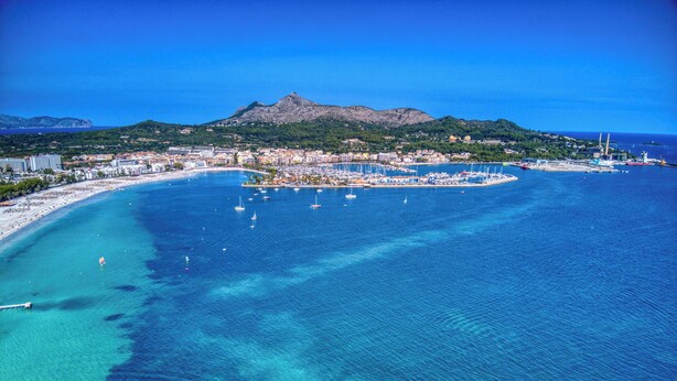 An aerial view of a coastal city with a marina, clear blue waters, and a mountain in the background. - Fantastic garden apartment directly on the dream beach of Puerto Alcudia (Alcúdia)