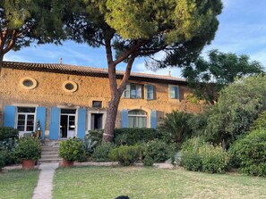 A rustic building with a tiled roof, a central entrance with steps, and a garden with various plants and a pathway leading to the entrance. - Apartment 70 m2 pool property, parking, sea 4 km (Sérignan)