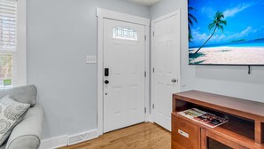 A room with a white door, a wooden cabinet with magazines, and a wall-mounted TV displaying a beach scene. - Sleeps 6 | Private Patio | Near Downtown CHS (North Charleston)