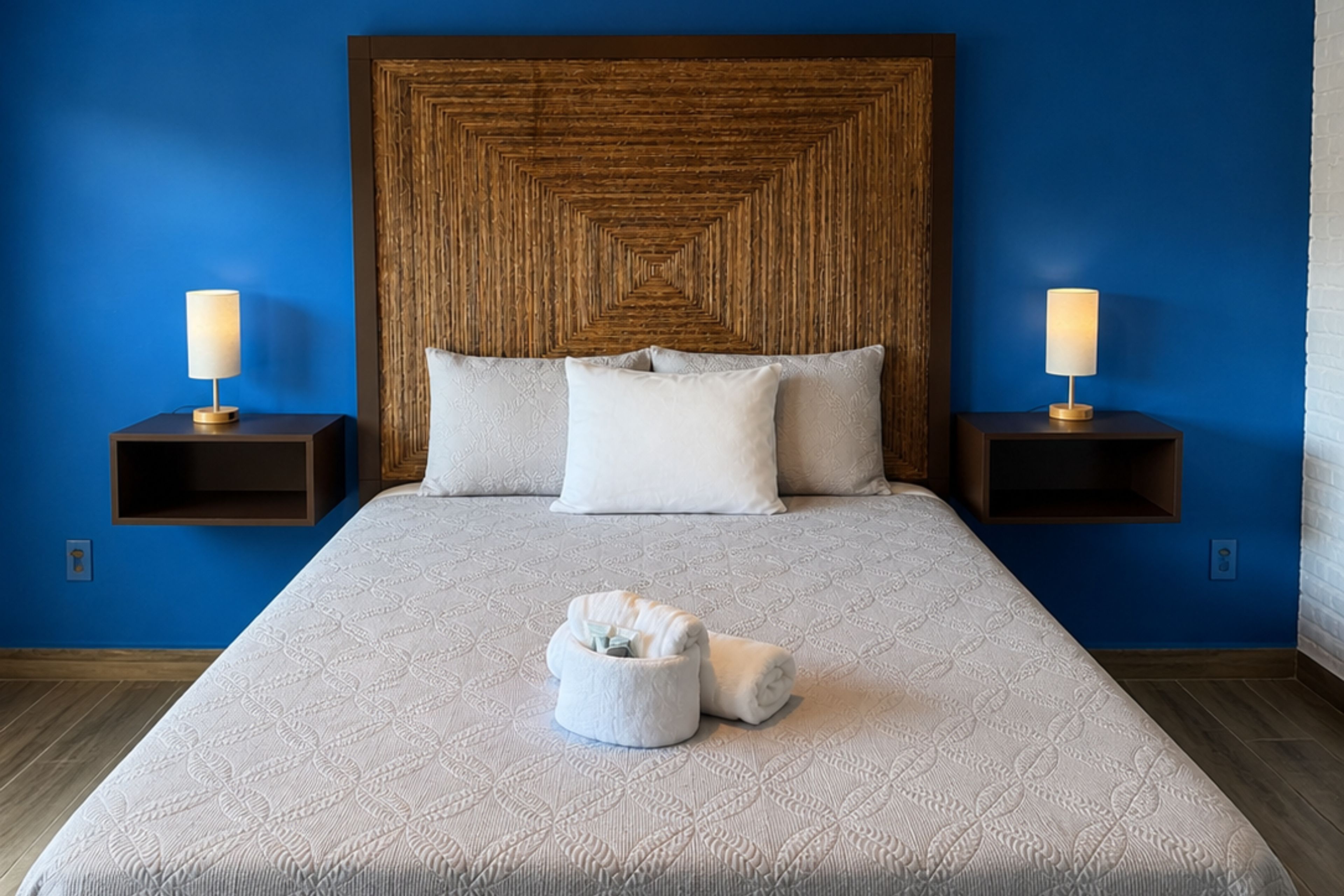Una habitación de hotel con una cama grande, dos mesitas de noche con lámparas, un cabecero con un diseño geométrico y una pequeña cesta blanca con toallas.