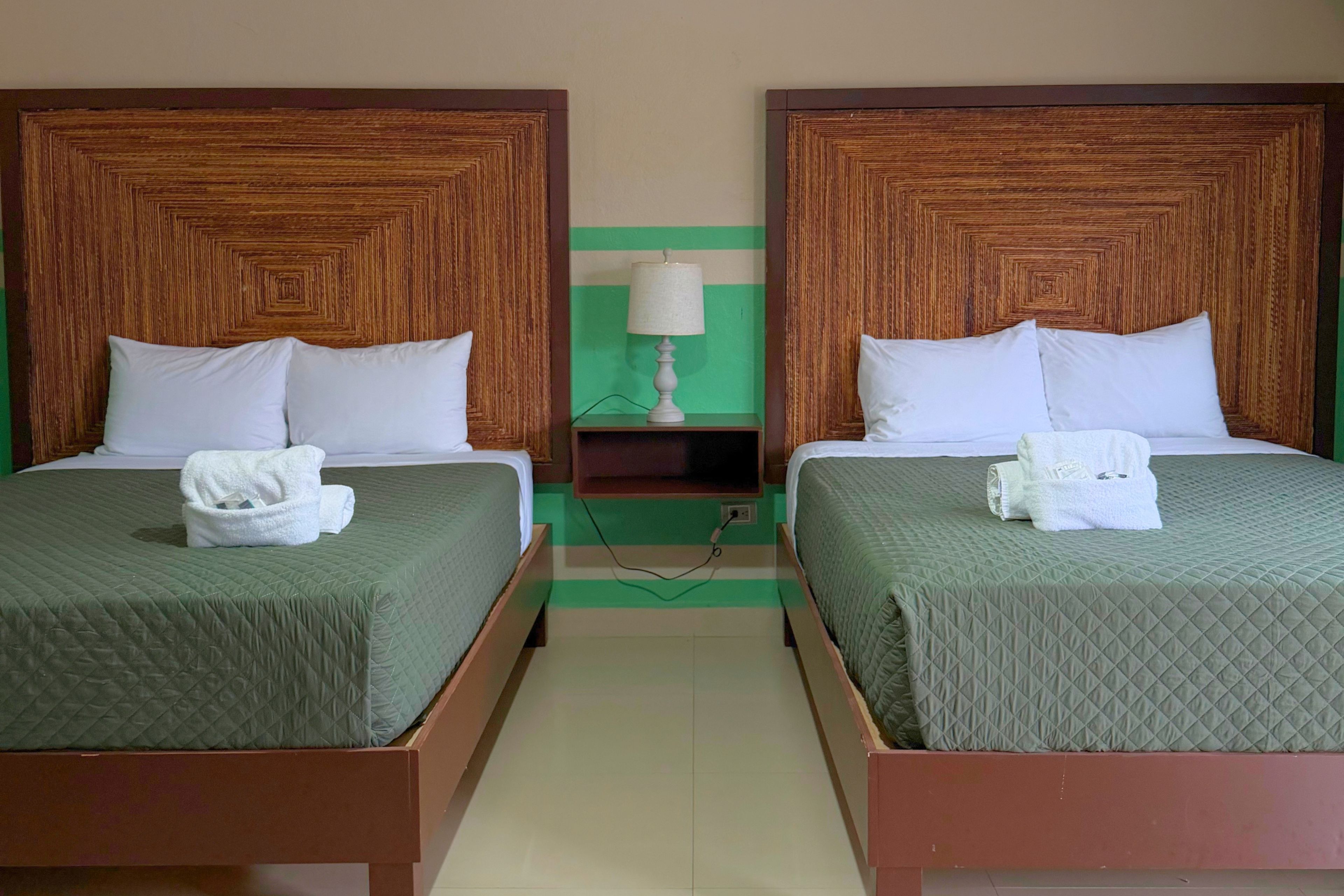 Una habitación de hotel con dos camas, cada una con cabecero de madera, edredón verde, almohadas blancas y una toalla blanca.