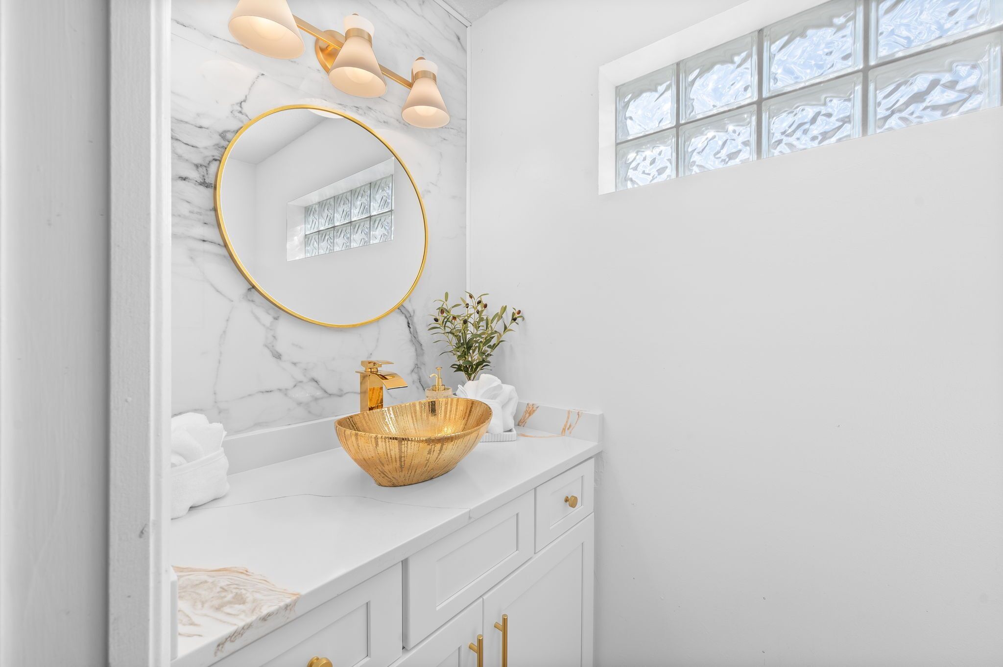 A bathroom with a marble vanity, a gold sink, a round mirror, and a window with frosted glass.